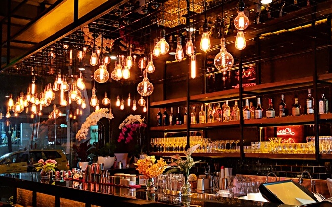 a lot of lightbulbs hanging overhead over a bar with lots of drinks